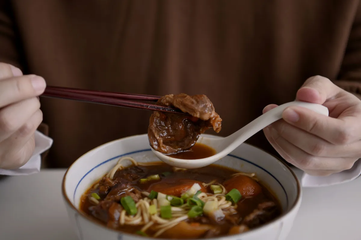 雙手用湯匙和筷子撈起一塊門前隱味燉煮牛肉,下面是一碗牛肉麵